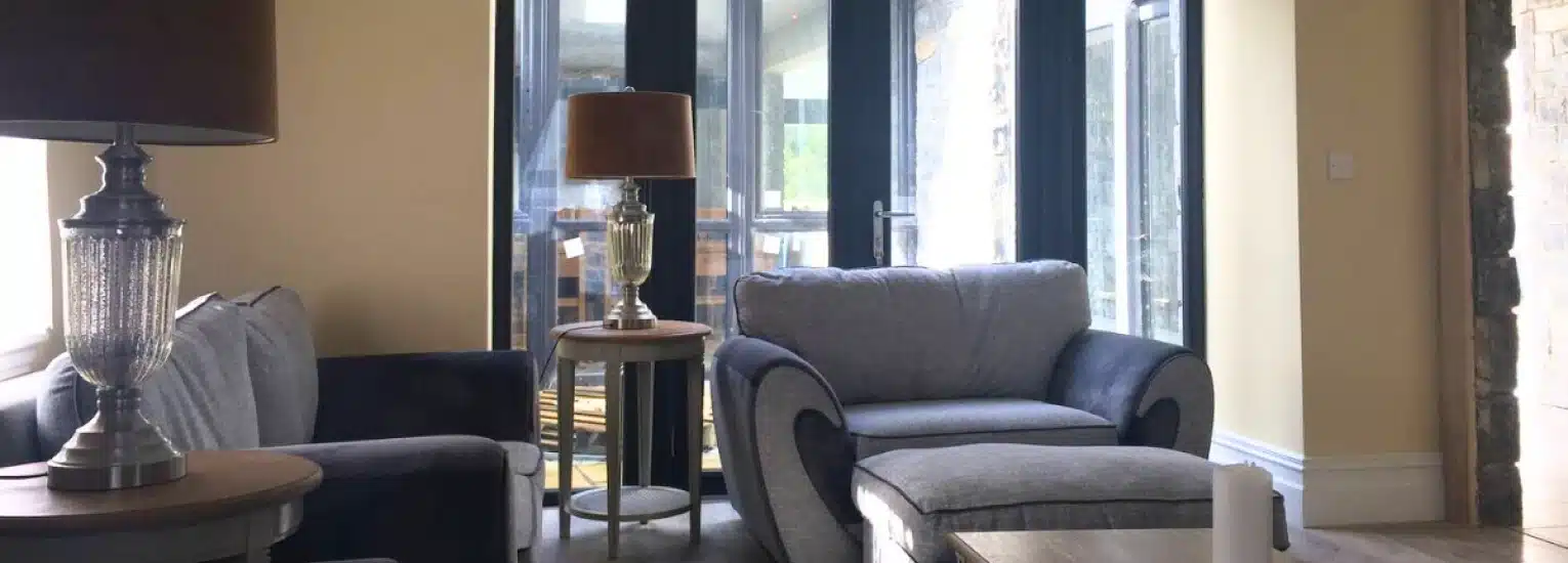 A cosy living room with two grey armchairs, a matching ottoman, two side tables with lamps, and large glass doors leading to a bright dining area. Cream-coloured walls and soft natural light create a welcoming atmosphere.