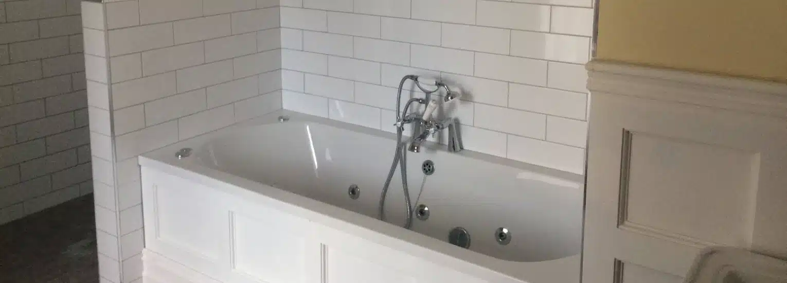 A white bath with built-in jets sits against a wall with white metro tiles and a silver tap, in a bathroom with yellow and white panelled walls.