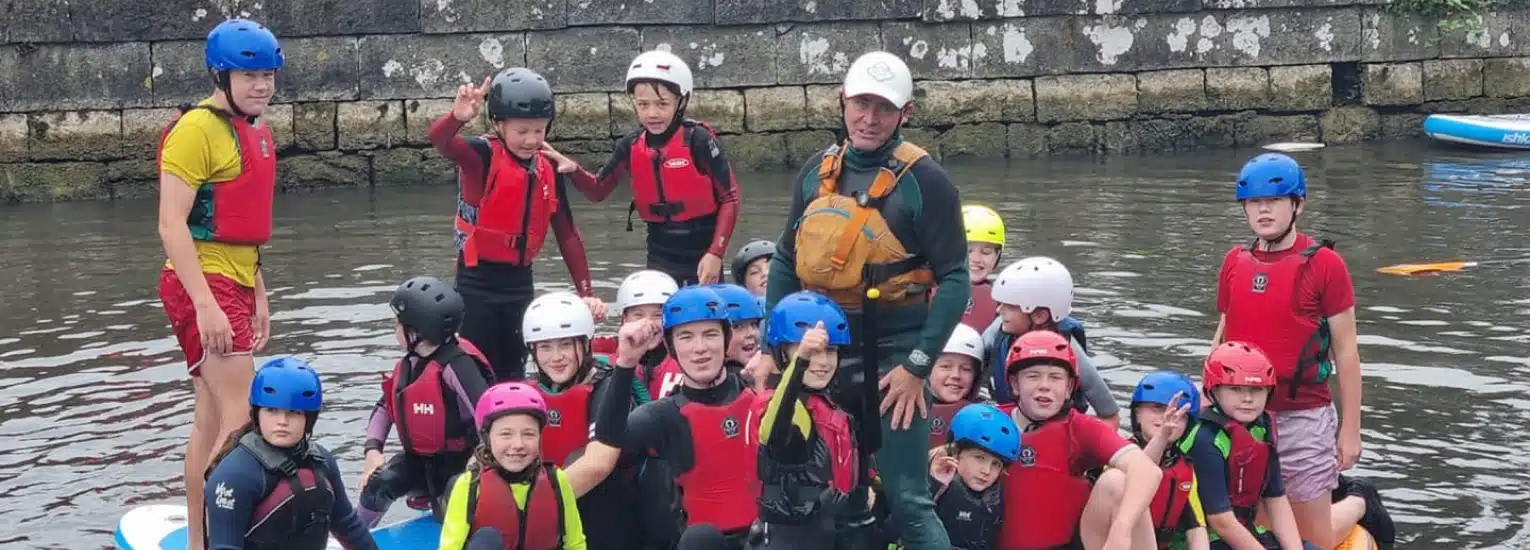 A group of children and an adult, all wearing helmets and life jackets, pose together in shallow water near a stone wall, some standing and some sitting on paddleboards, smiling and making peace signs.
