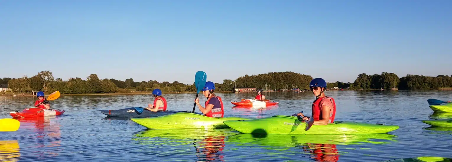 Several people wearing lifejackets and helmets paddle colourful kayaks on a calm lake under a clear blue sky, with green trees and distant buildings visible along the shore.