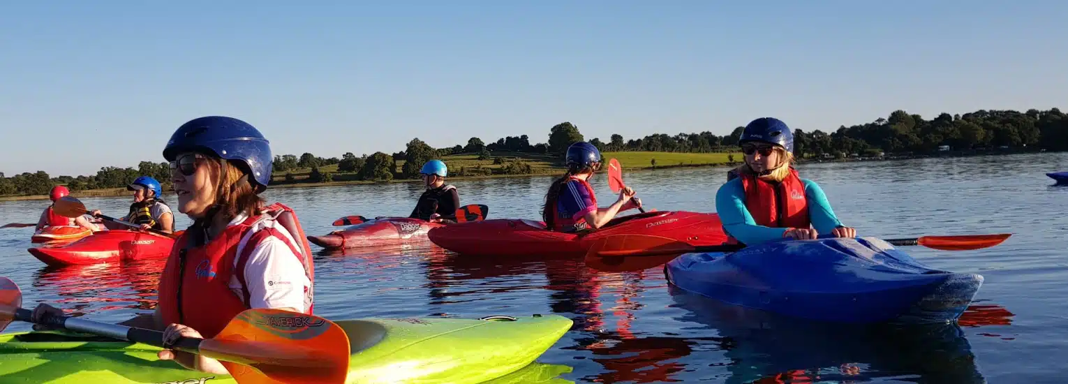 Several people wearing helmets and life jackets paddle colourful kayaks on a calm lake, with trees and grassy hills visible in the background under a clear blue sky.