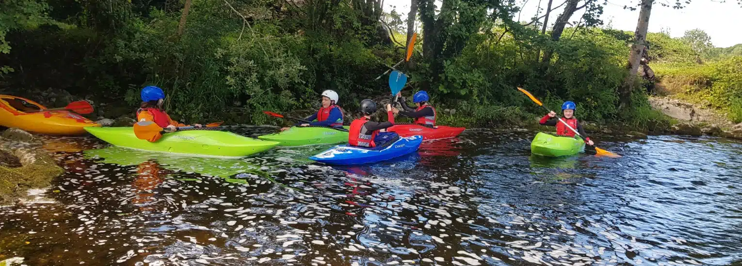 A group of people wearing helmets and life jackets paddle colourful kayaks on a calm river near a leafy, tree-lined bank.