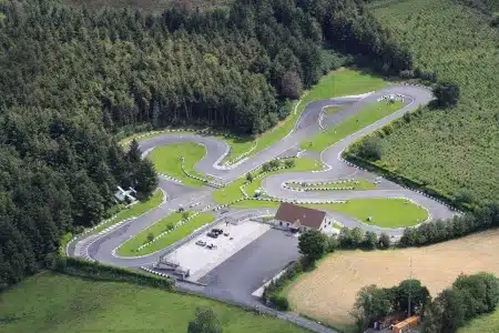 Aerial view of a winding go-kart track surrounded by trees and fields, with a building and car park near the entrance and green grassy areas between the track bends.