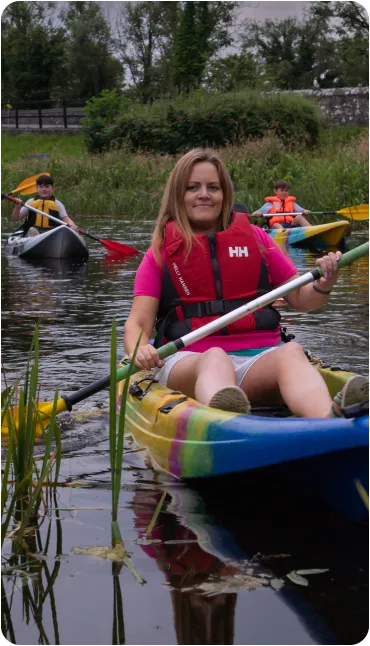 A woman wearing a pink shirt and red life jacket paddles a blue kayak on a calm river, followed by two children in kayaks. Lush green reeds and trees line the water’s edge.