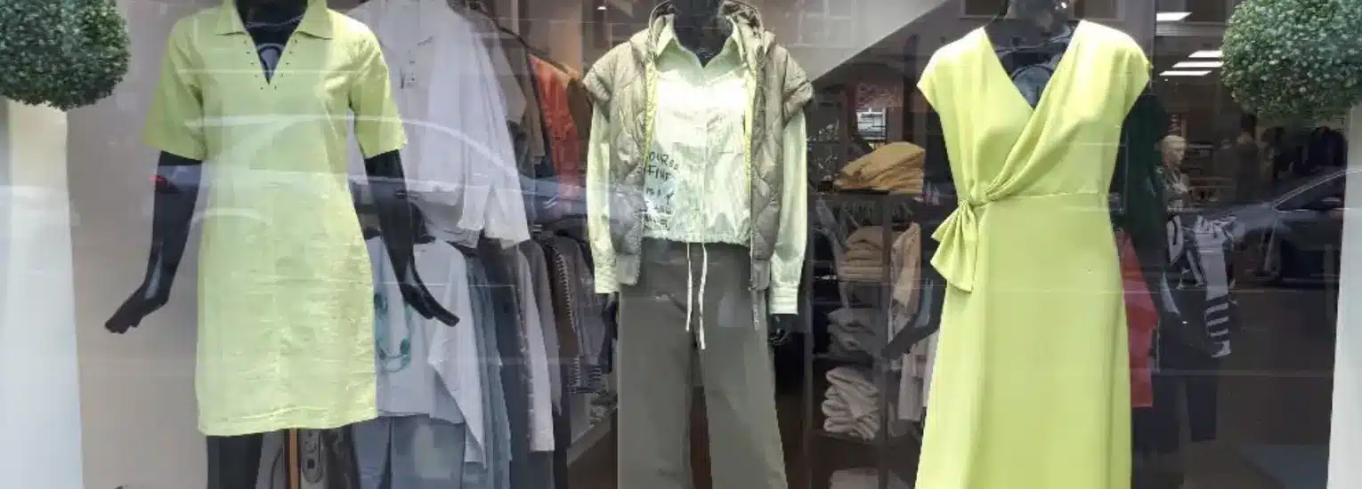 Shop window display with three mannequins wearing spring outfits: a light green short-sleeve dress, a light green shirt with a green jacket and khaki trousers, and a sleeveless yellow wrap dress.