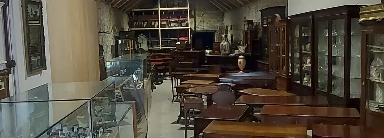 A room filled with antique wooden furniture, glass display cabinets, and shelves holding various collectables, all arranged neatly against stone walls under a sloping wooden ceiling.