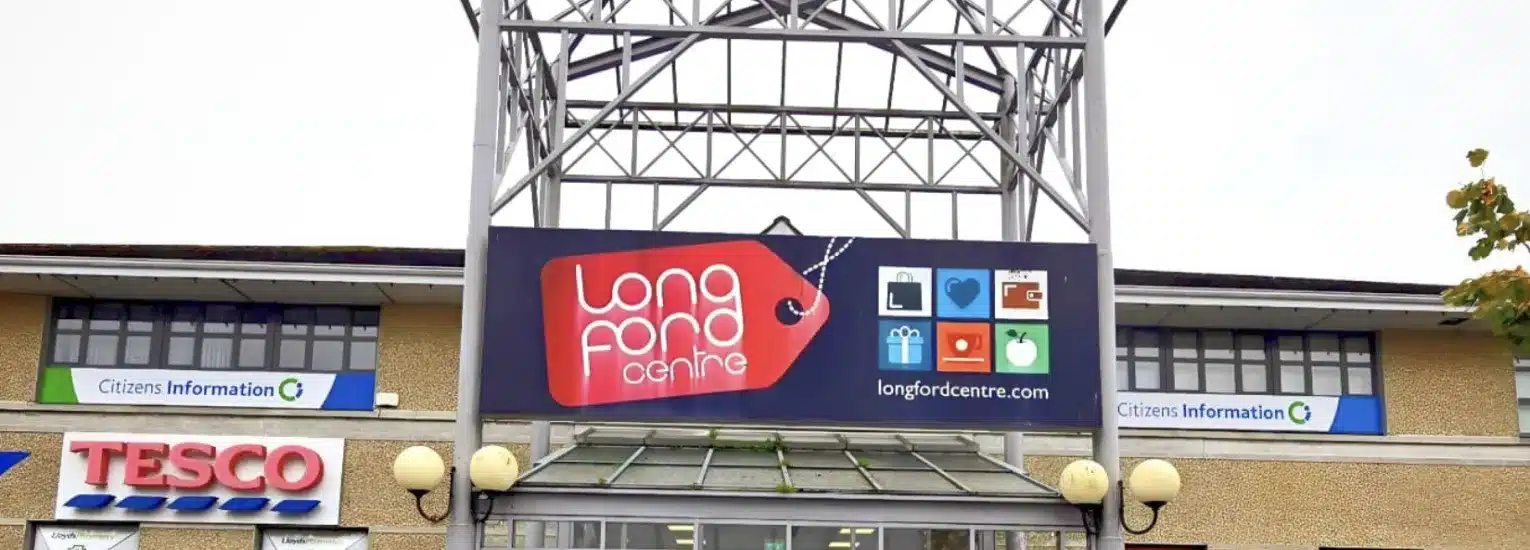 Front view of the Longford Centre shopping mall entrance, featuring a large sign with icons and the centre’s name above glass doors, with Tesco and Citizens Information signs visible below.