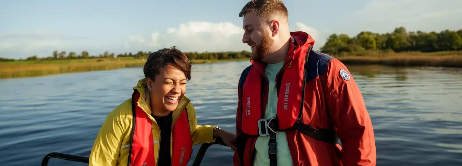 Two people wearing life jackets and jackets laugh together whilst standing on a boat on a calm lake, with green trees and blue sky in the background.