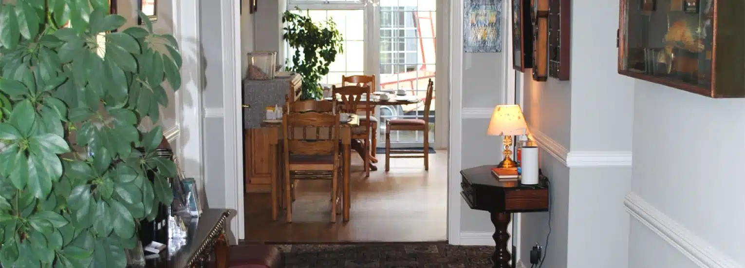 A bright hallway with plants and framed pictures on the walls leads to a dining area with wooden chairs and a table. A small table with a lamp stands to the right, and a window lets in natural light at the far end.
