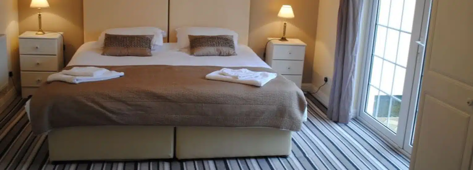 A neatly made double bed with brown and white bedding, two bedside tables with lamps, towels folded at the foot of the bed, striped carpet, and a glass door letting in natural light.