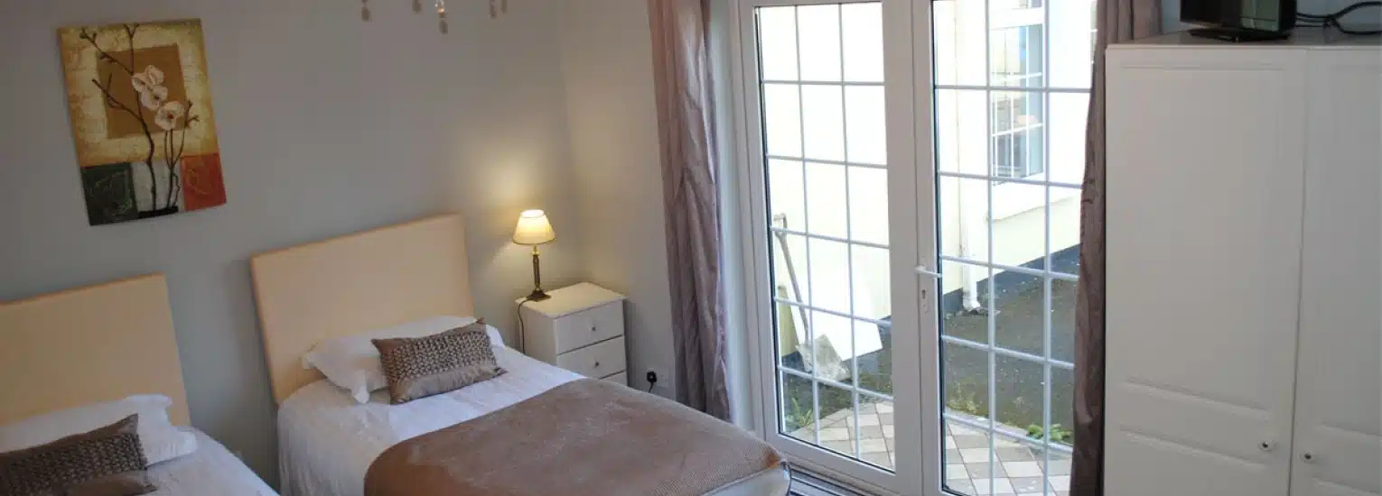 A bright bedroom with two single beds, a bedside table with a lamp, a white wardrobe, a floral painting on the wall, and large glass doors leading to a patio outside.
