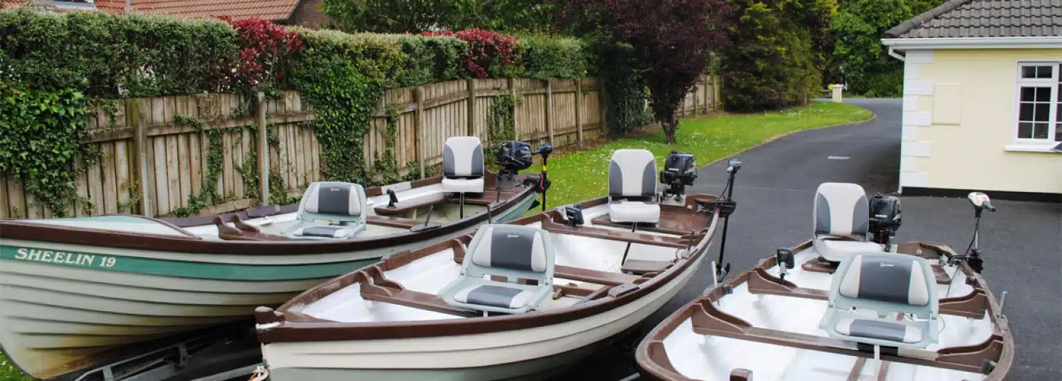 Three small fishing boats with motors are parked on a paved drive next to a cream-coloured house, with a wooden fence and green bushes in the background.