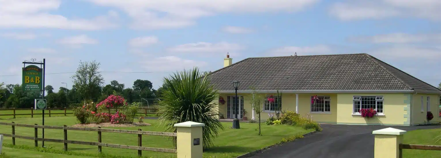 A single-storey yellow bed and breakfast building with a grey tiled roof, surrounded by a well-kept lawn, flowers, a driveway, and a wooden fence; a signpost for the B&B stands at the entrance.