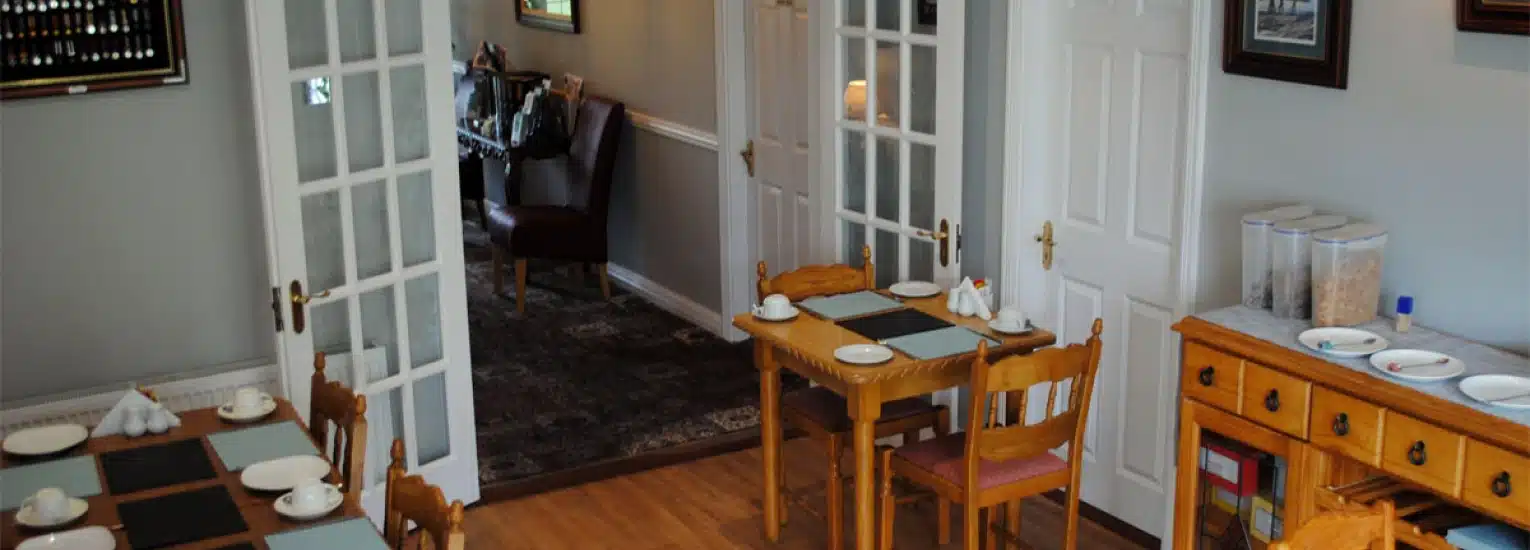 Cosy dining area with small wooden tables set for breakfast, each with cups, plates, and placemats. A wooden sideboard with cereal containers is against the wall, and a lounge area is visible through open glass doors.