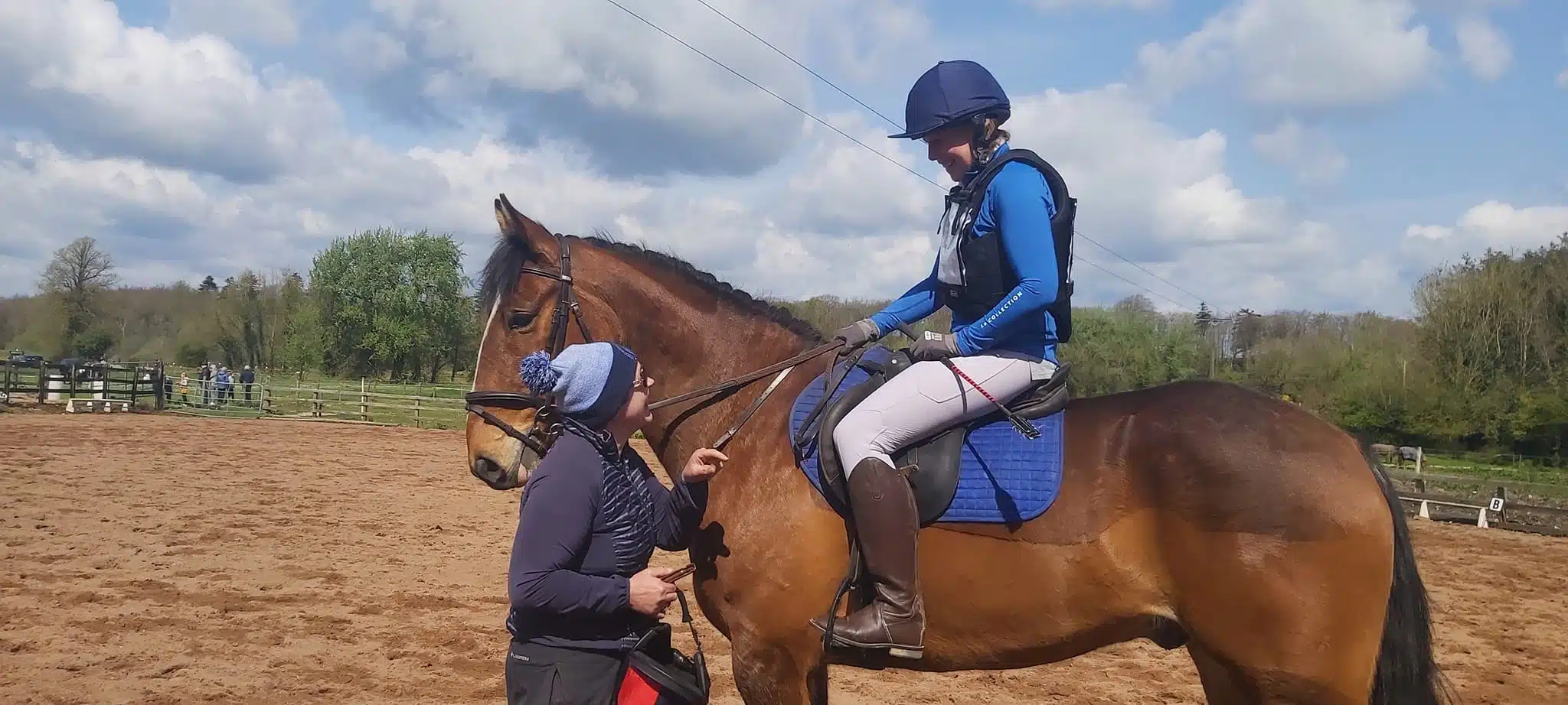 A person on horseback in riding kit listens to an instructor who is standing beside the horse in a sandy outdoor arena, with trees and a cloudy blue sky in the background.