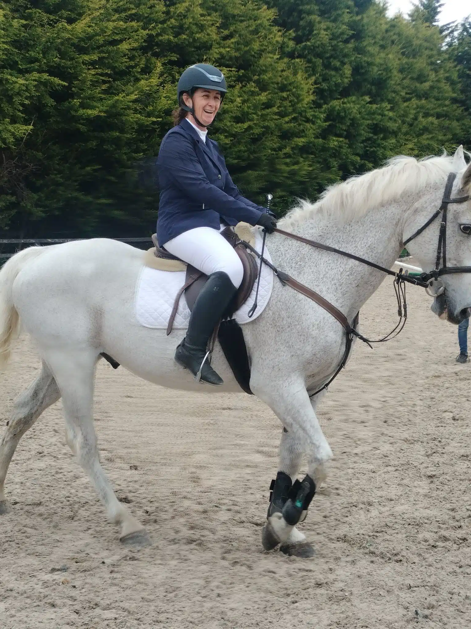 A person wearing riding kit and a helmet is happily riding a white horse with a white numnah in a sandy outdoor arena, with green trees in the background.