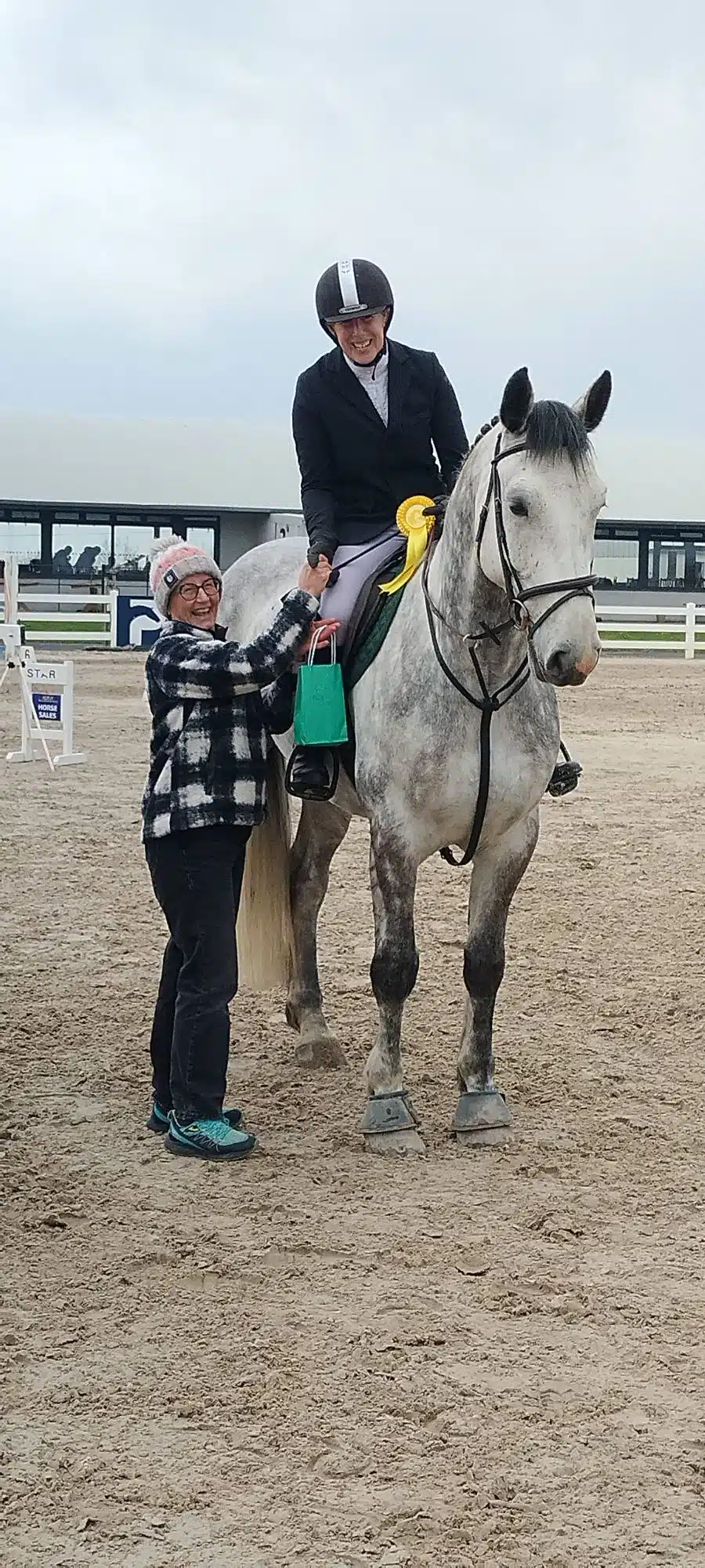 A smiling person on a grey horse wearing a riding helmet and holding a yellow rosette, while another person stands beside them holding the horse’s tail and a green gift bag. The scene is at an outdoor equestrian event.