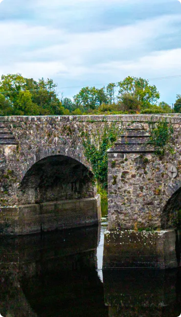 A stone bridge with large arches crosses over a calm river, surrounded by greenery and trees under a cloudy sky.