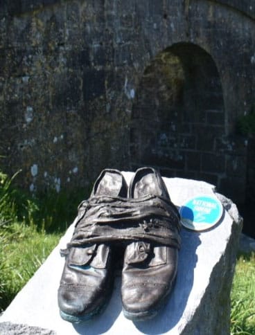 A pair of worn, dark shoes wrapped with cloth sit on a stone slab outdoors, with an arched stone bridge and grass in the blurred background. A round blue badge is next to the shoes.