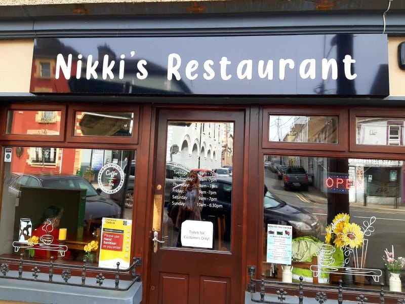 The exterior of Nikki’s Restaurant with large windows, a dark blue sign with white lettering, visible flowers and decorations inside, and an "OPEN" sign on the door. Reflections of nearby buildings appear in the glass.