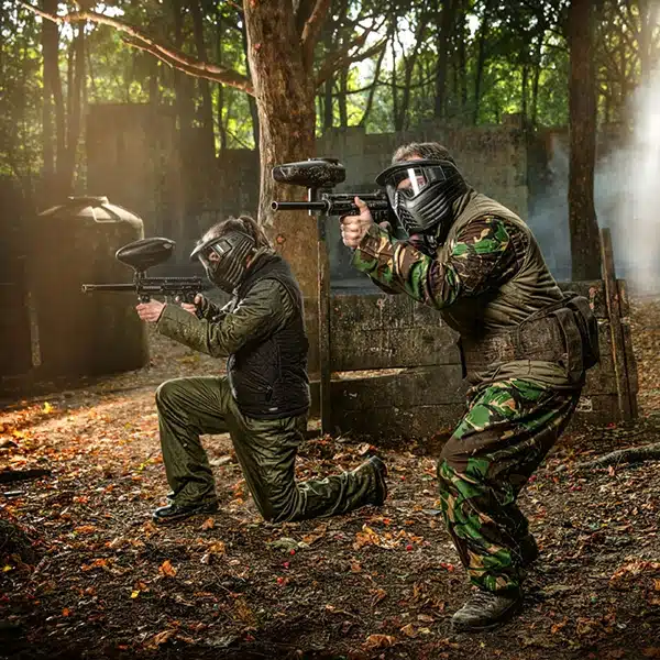 Two people wearing protective masks and camouflage clothing play paintball in a wooded outdoor area. One is kneeling and aiming a paintball gun, while the other stands behind, also aiming, with sunlight filtering through the trees.