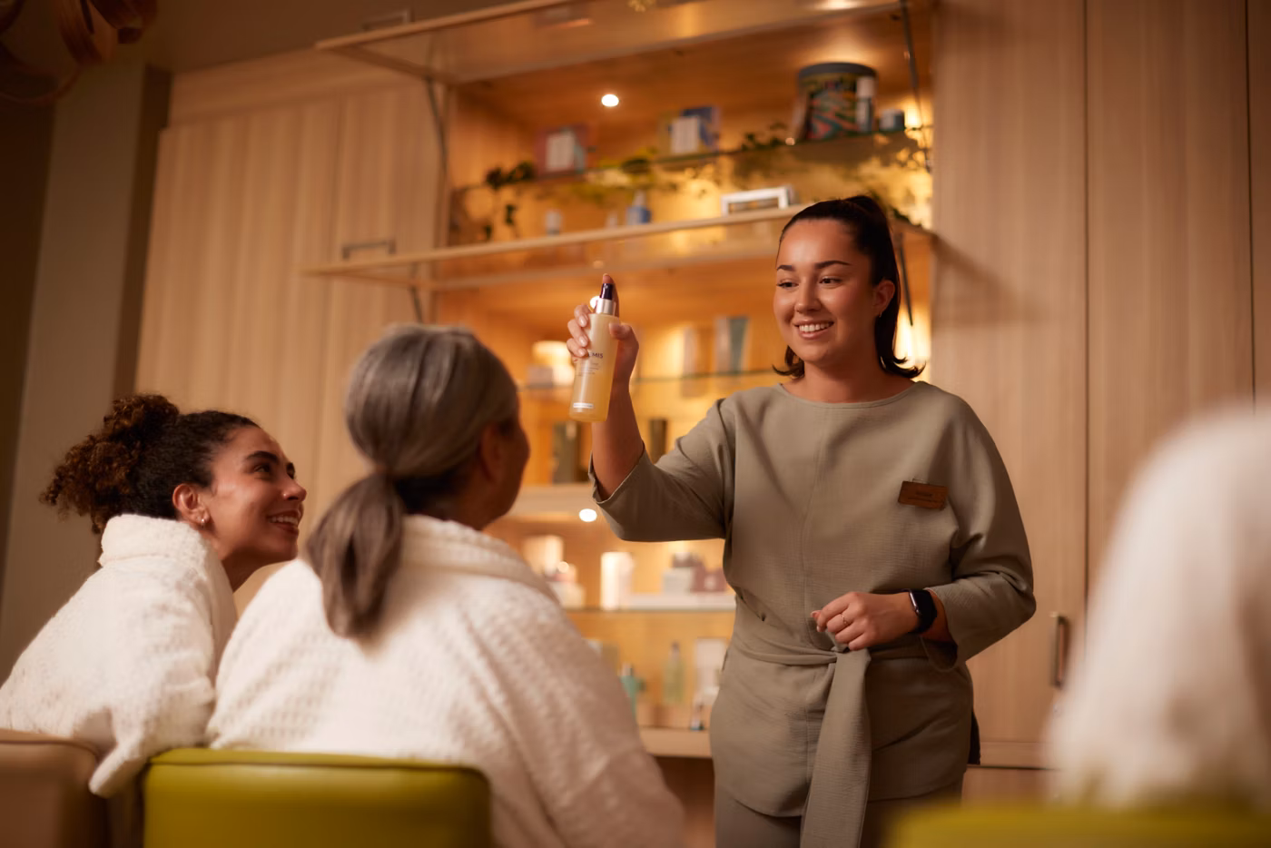 A spa employee in a uniform demonstrates a skincare product to two women wearing white dressing gowns, one with grey hair and one with curly hair, in a warmly lit, modern spa room with shelves in the background.