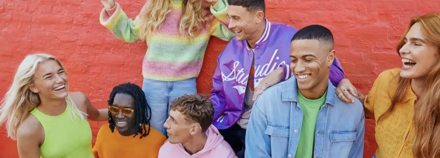 A group of seven young adults laughing and smiling together in front of a bright red wall, dressed in colourful, casual clothing.