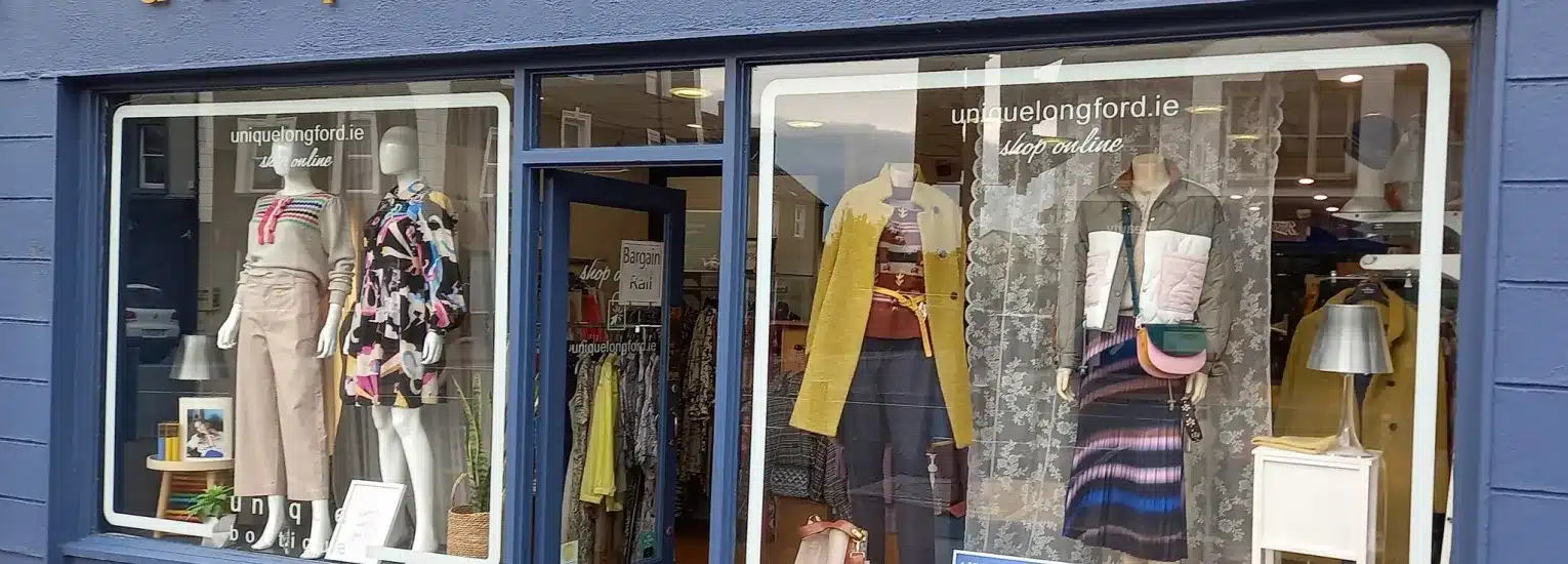A clothing shop window display featuring mannequins dressed in colourful outfits, including skirts, trousers, tops, and jackets. The shop’s name and website appear on the window above the mannequins.