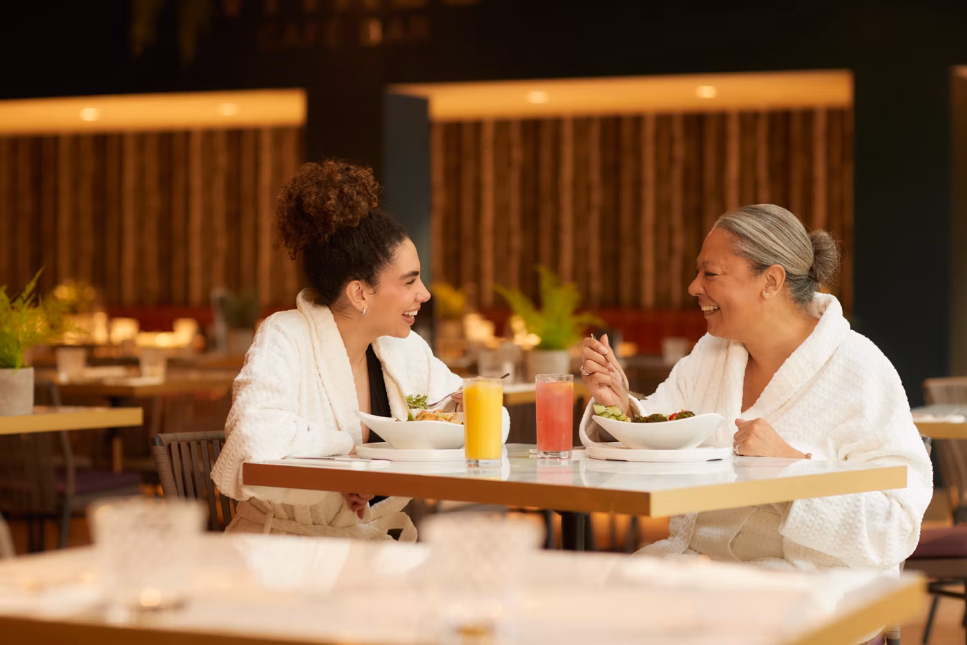 Two women wearing white dressing gowns sit at a restaurant table, smiling and enjoying a meal together. Drinks, including a glass of orange juice and a pink drink, are on the table, with a warm, relaxed atmosphere surrounding them.
