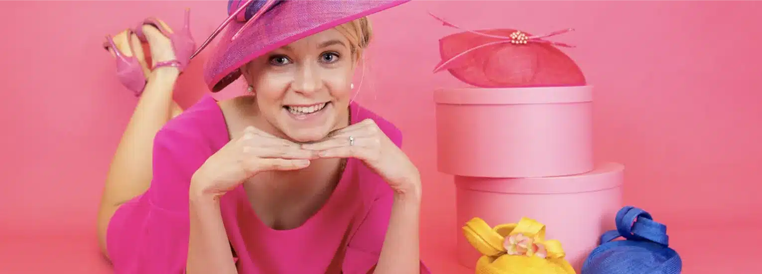 A person in a bright pink dress and wide-brimmed hat smiles whilst lying on the floor with hands under their chin. Pink hat boxes and colourful hats in yellow and blue are arranged nearby against a pink background.
