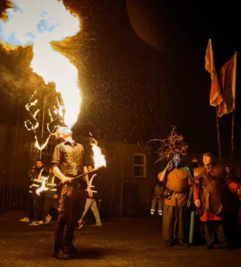 A performer breathes fire into the night sky with a flaming torch, as people in costumes—including one with blue face paint and another holding flags—watch in the background, adding vibrant energy to a memorable Sustainable Tourism Longford event.