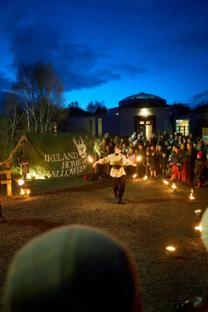 A performer twirls flaming batons before a crowd at night in Longford, with a large green banner reading “Ireland Home of Hallowe’en” in the background—an exciting celebration promoting sustainable tourism under a dark blue sky.