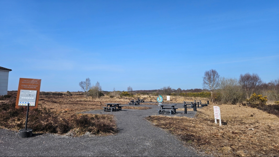 A paved path leads to several picnic tables and benches in an open, dry grassland area under a clear blue sky. Signs are posted near the path and on the left side of the image. Sparse bushes and trees surround the area.