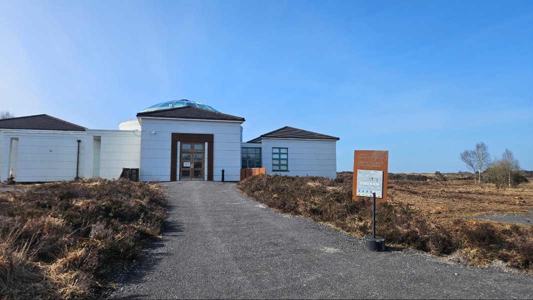 A small white building with a glass dome on the roof sits at the end of a paved path, surrounded by dry grass and an open landscape under a clear blue sky. A brown sign stands beside the path near the entrance.