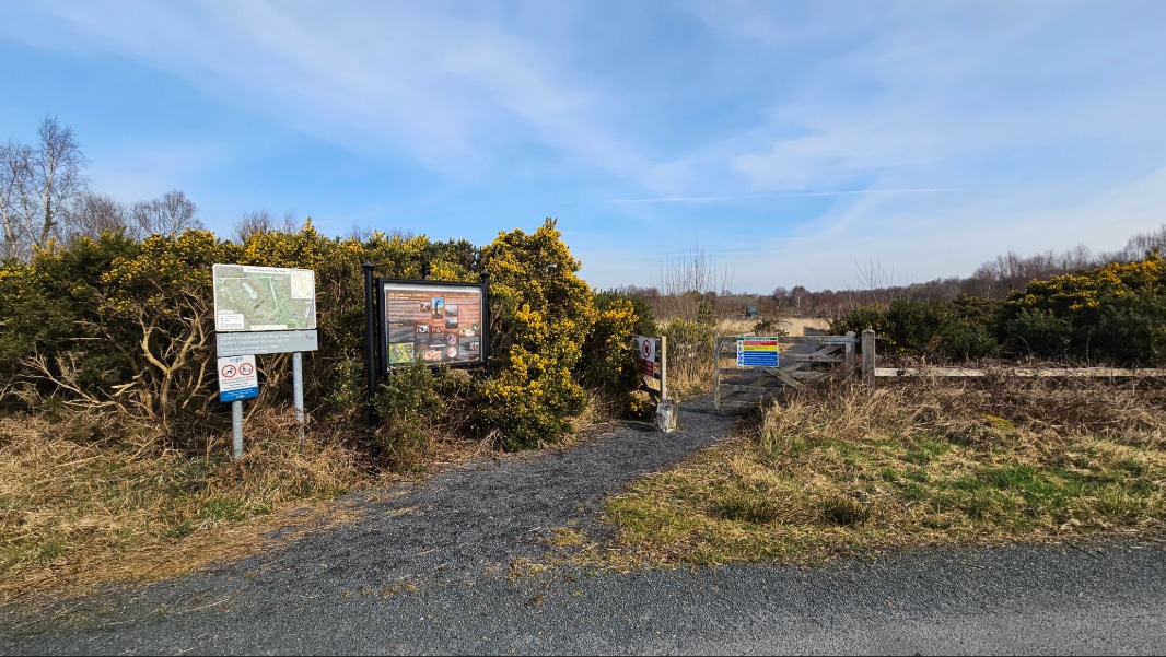 A rural footpath entrance with information boards and signs, bordered by bushes and grassy areas. A dog stands on the path near the signs; trees and open fields are visible in the background under a blue sky.