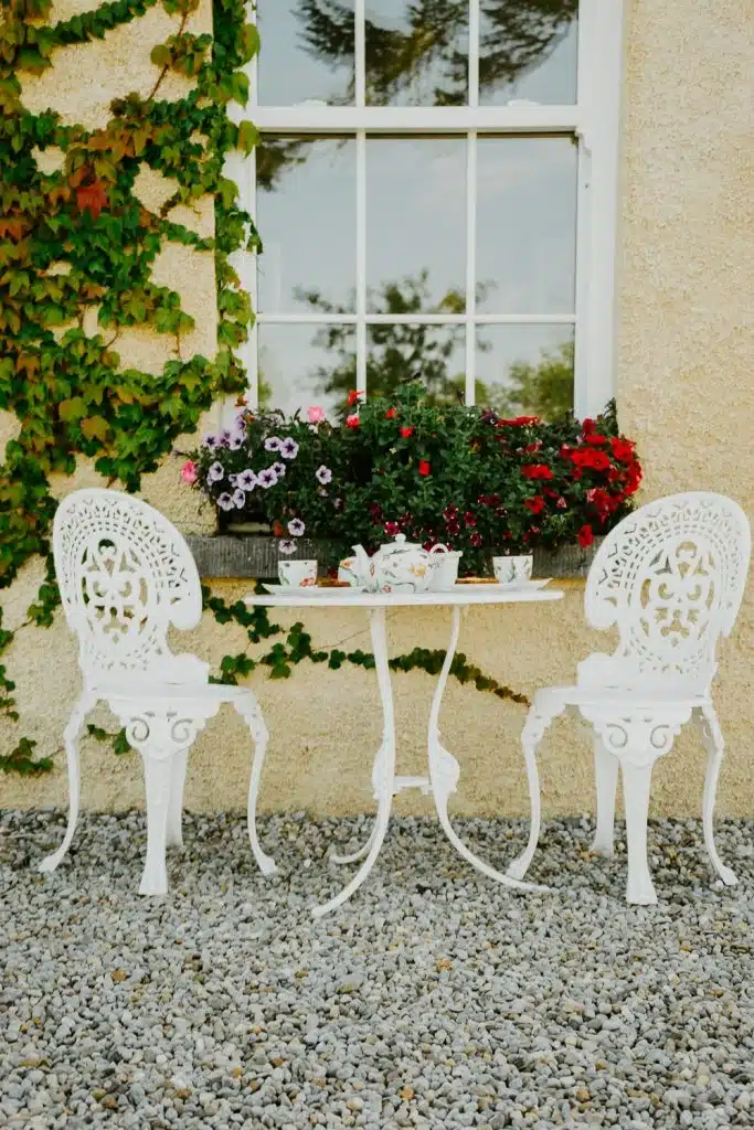 A small white metal table with two ornate matching chairs sits on gravel outside a window, surrounded by green ivy and colourful flowers in a window box. Tea cups and a teapot are set on the table.