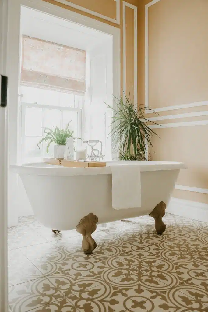 A white clawfoot bath with a towel draped over the side sits in a stylish bathroom with patterned tiled flooring, tan and white walls, large window, potted plants, and a tray holding bath items.