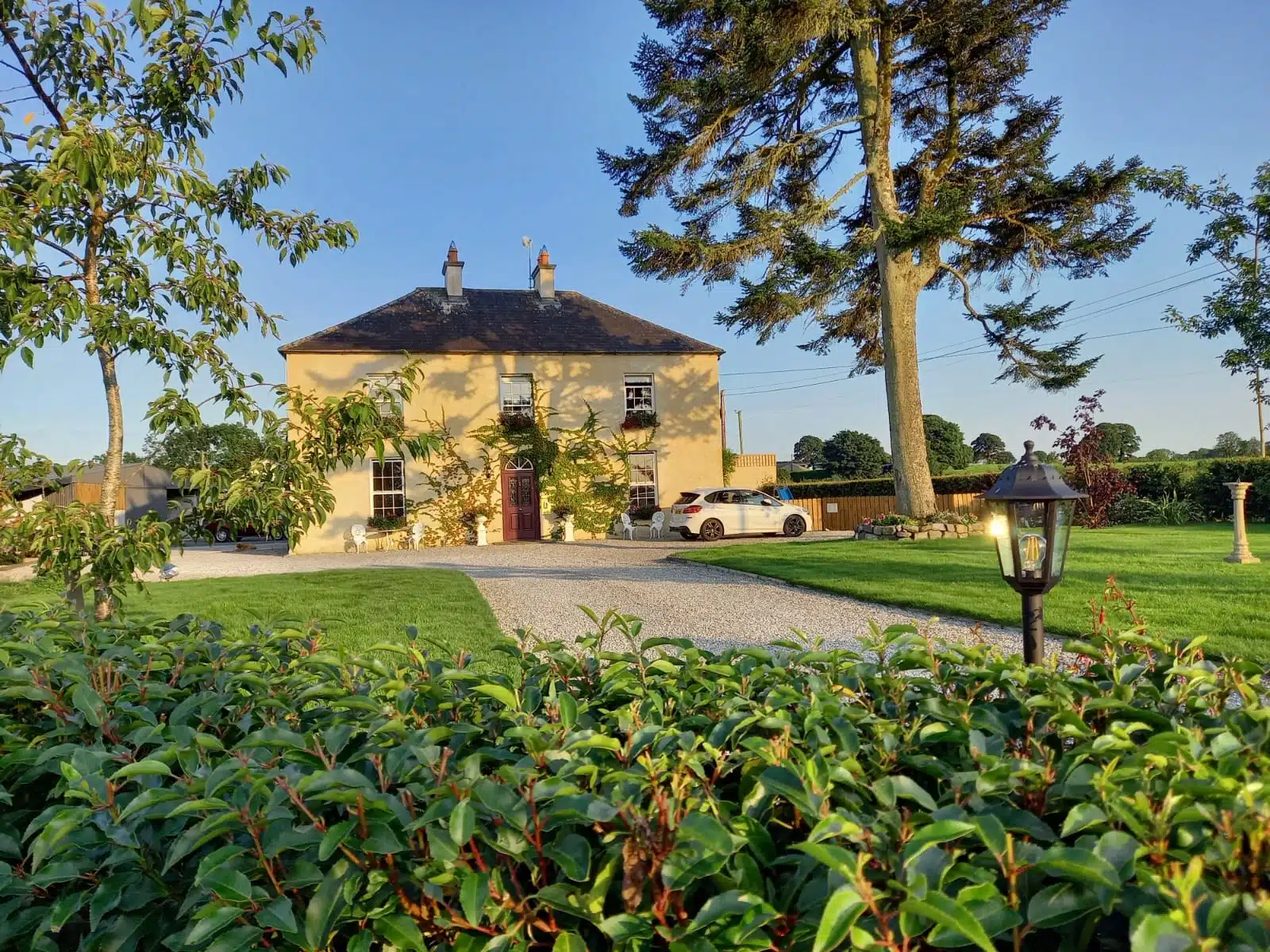A large yellow two-storey house with a dark roof stands in a manicured garden with green grass, tall trees, a gravel drive, and a white car parked beside the house on a sunny day. A lamp and bushes are in the foreground.