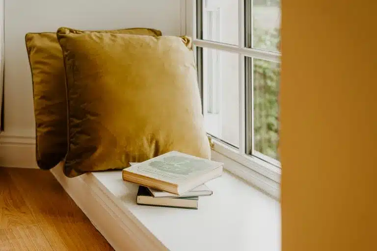A cosy window seat with mustard yellow cushions and three stacked books, one open, next to a large window letting in natural light.