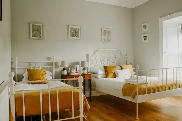 Two white metal-framed beds with mustard yellow bedding and pillows in a cosy, light-filled bedroom with wooden floors. A small wooden table with lamps and decor sits between the beds. Framed art hangs on the pale walls.