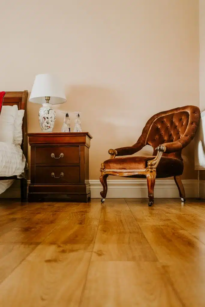 A cosy bedroom corner with a vintage brown upholstered armchair, a wooden bedside table holding a white lamp and glass bottles, all set on a polished wooden floor against a beige wall.