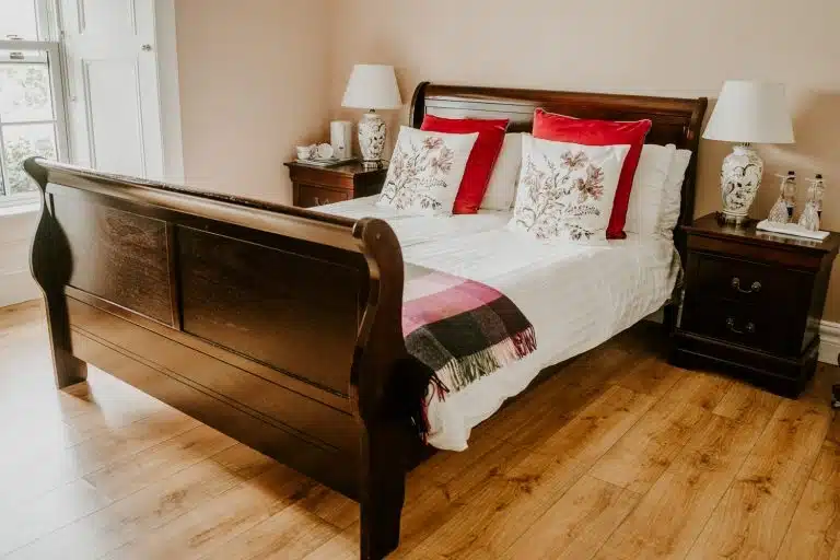 A cosy bedroom with a wooden sleigh bed, white linen, floral and red accent cushions, a checked throw, and matching wooden bedside tables with lamps and decorative items on a light wood floor.