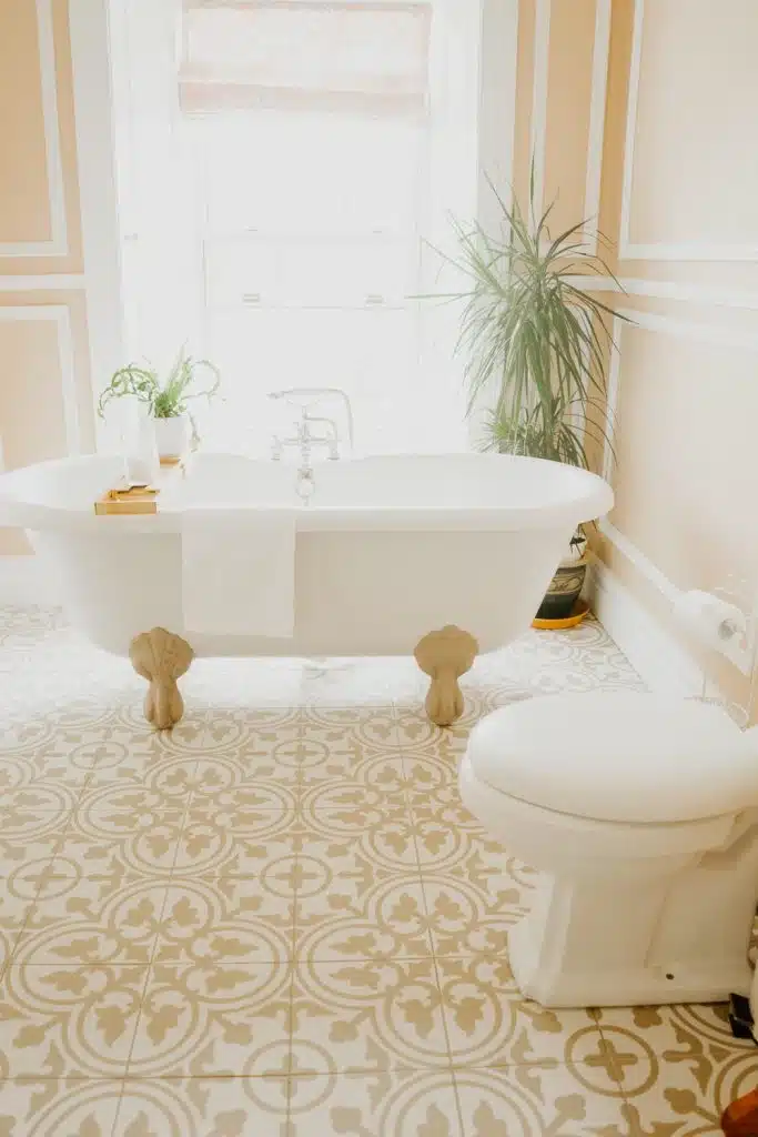 A bright bathroom with patterned tiled floor, a white clawfoot bath with a towel draped over the side, potted plants beside the bath, beige walls with white trim, and a white toilet in the foreground.