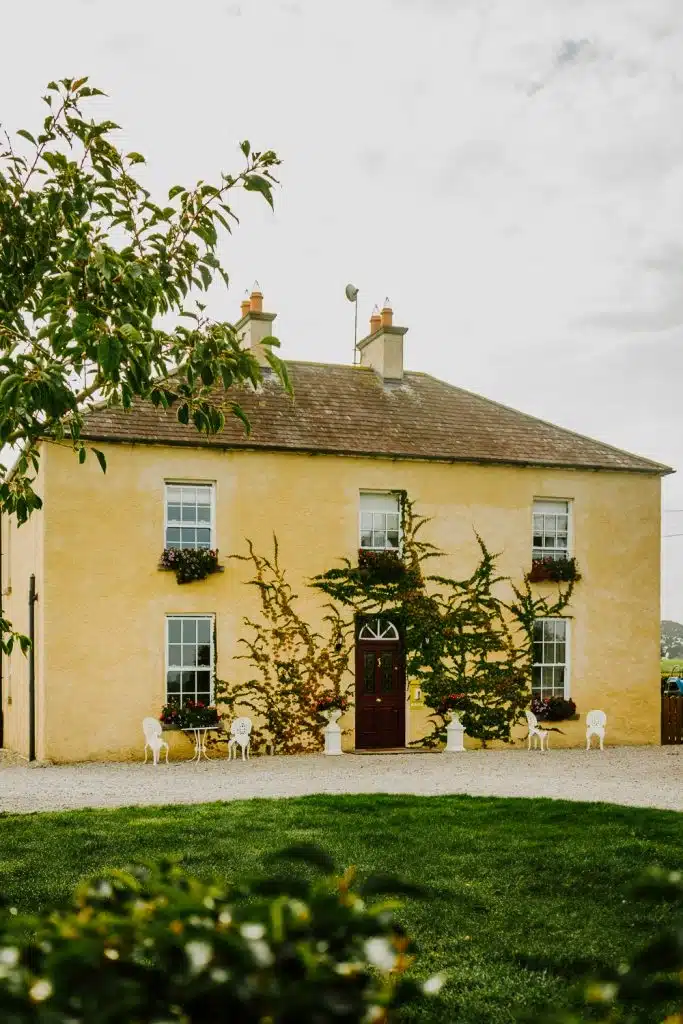 A two-storey yellow house with ivy climbing its walls, white-framed windows, flower boxes, a wooden door, and white outdoor chairs on a gravel path, surrounded by green grass and trees.