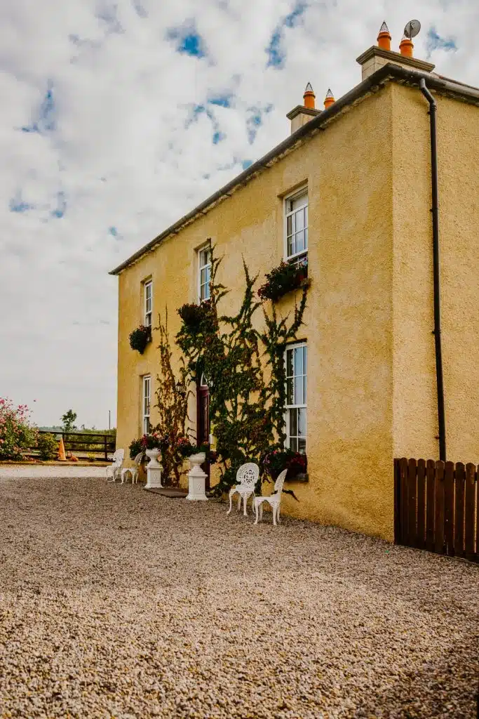 A two-storey yellow house with ivy climbing the walls, white-trimmed windows, flower boxes, and white chairs on a gravelled garden under a partly cloudy sky.