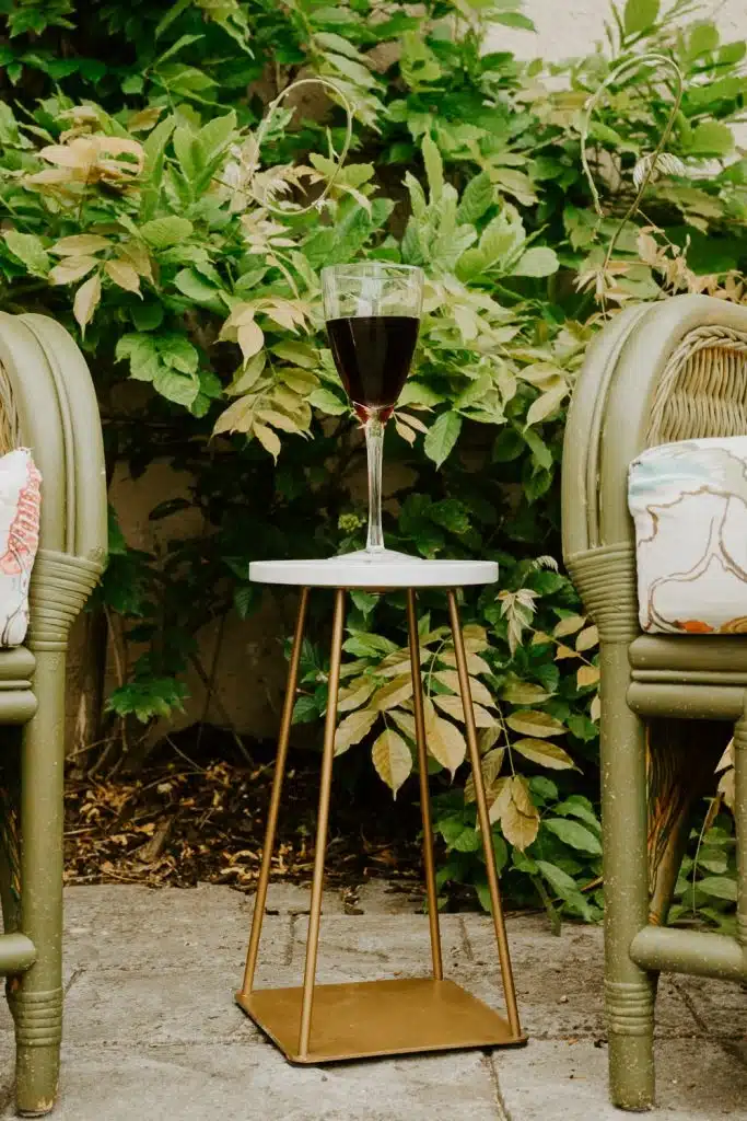 A glass of red wine sits on a small, modern side table between two green cushioned chairs, with lush green foliage in the background.