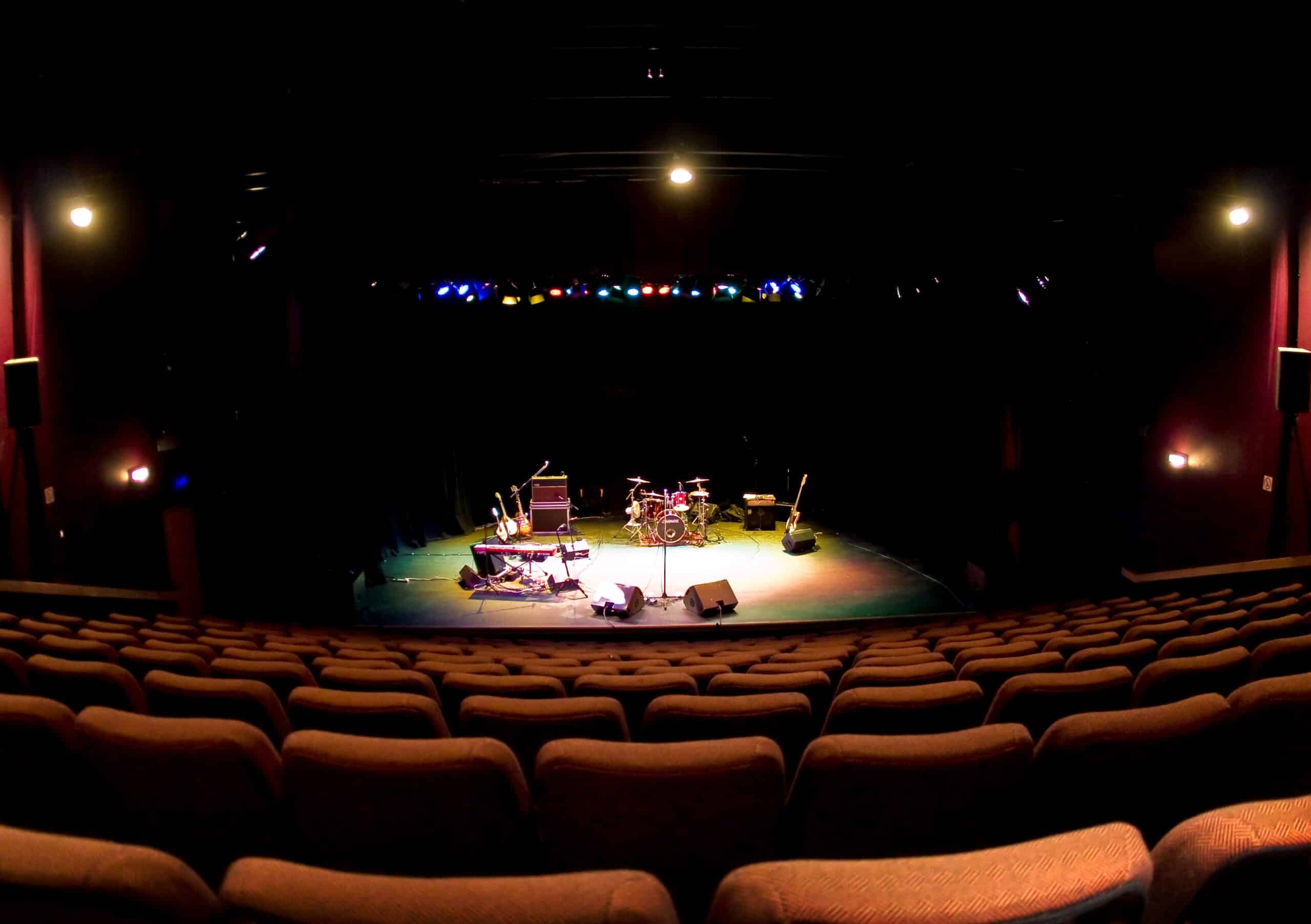 An empty concert hall stage with instruments set up and colourful lights above, viewed from the audience seating.