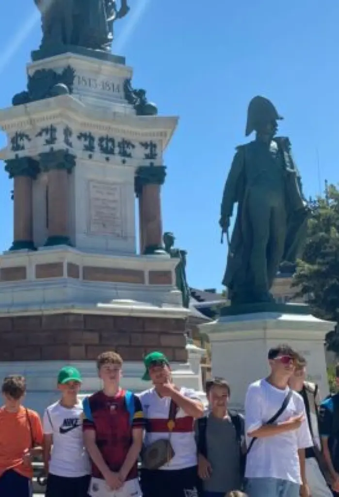 A group of boys stand in front of a large monument featuring a statue of an historical figure in uniform. Some of the boys wear hats, and it is a sunny day with trees and blue sky in the background.