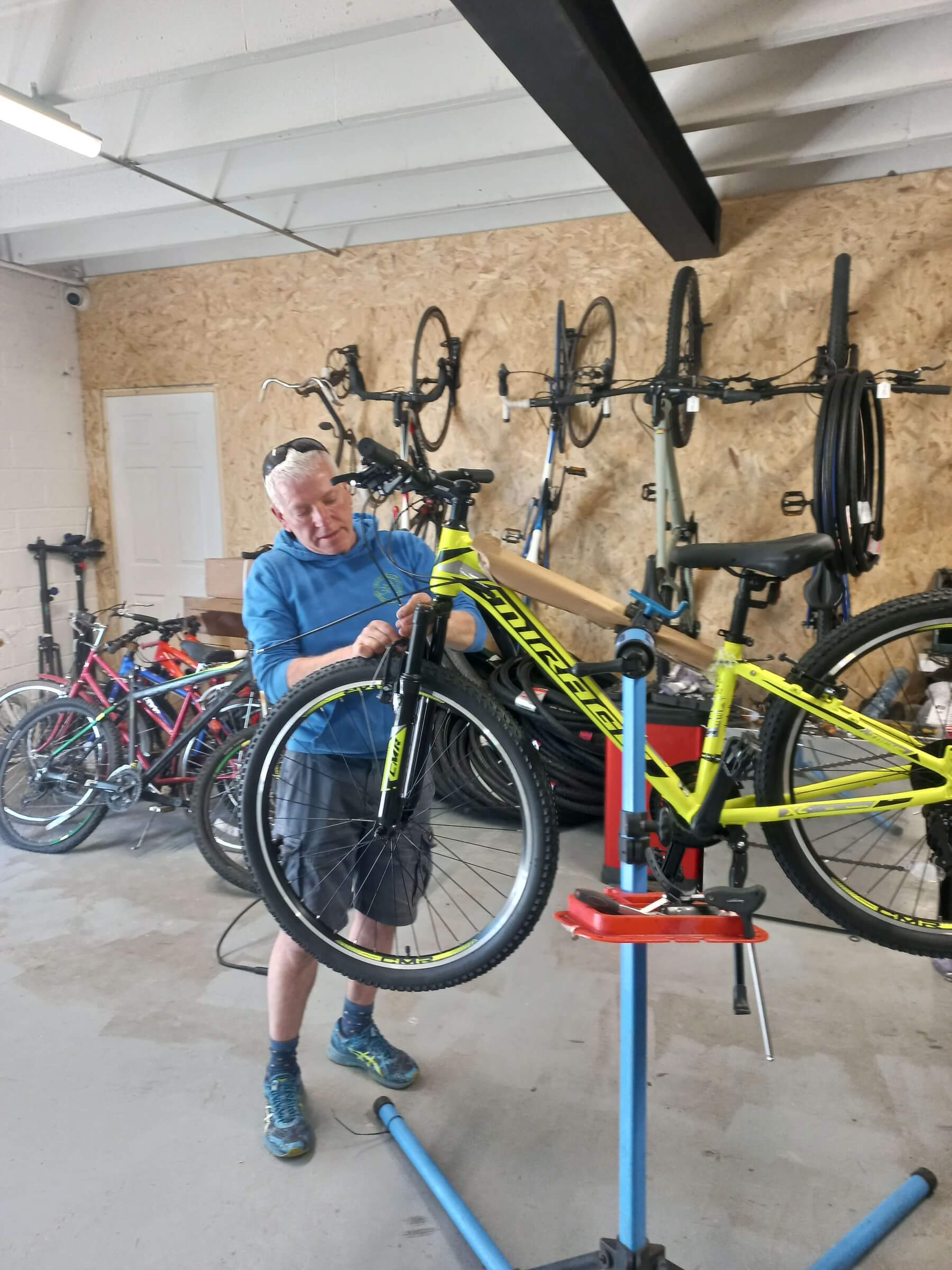 An older man in a blue shirt repairs a yellow bicycle on a stand in a workshop with bikes and wheels hanging on the wall behind him.