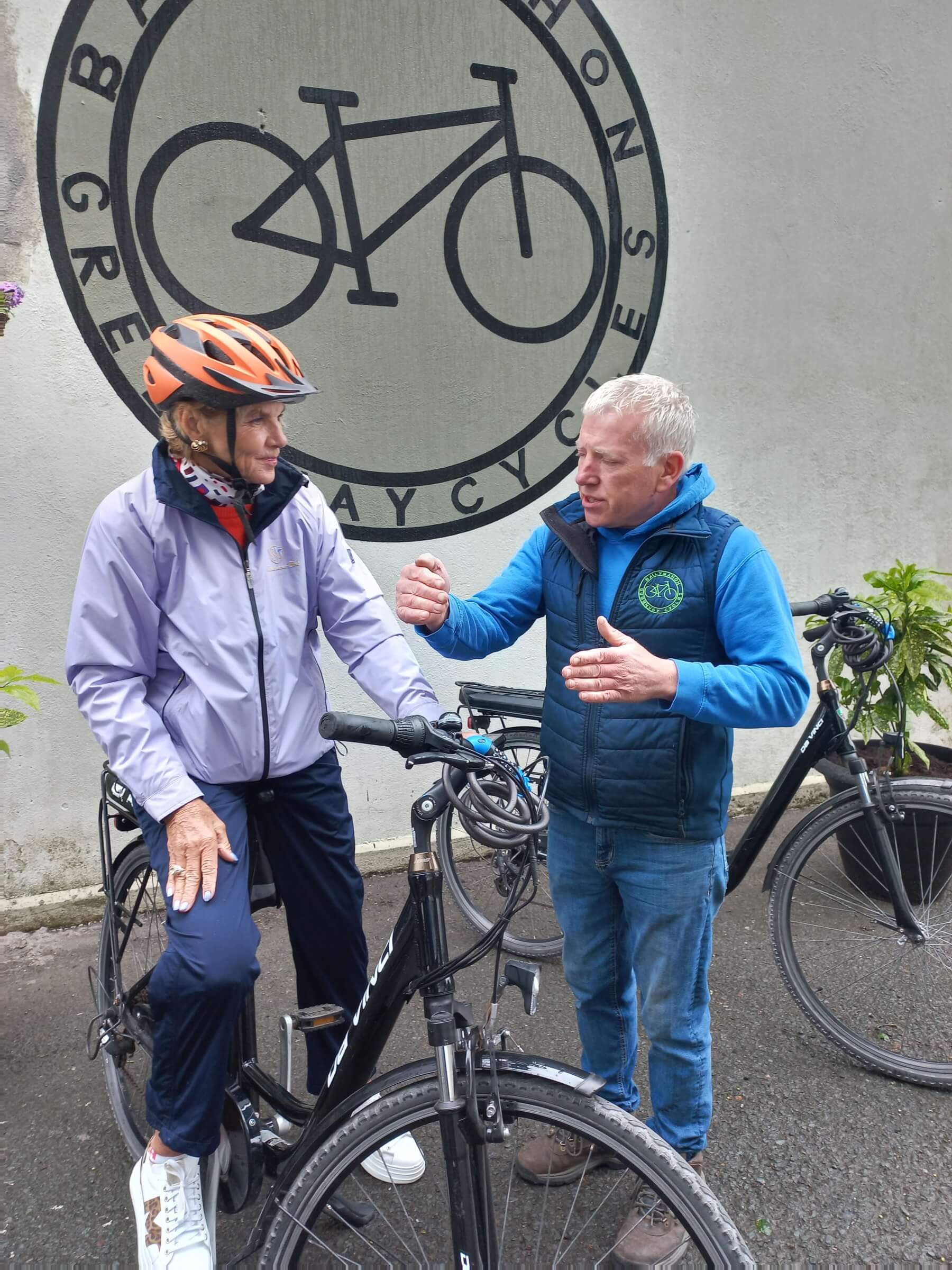 An older person wearing a helmet sits on a bicycle, whilst another person stands beside them gesturing and talking. They are outside in front of a wall with a large bicycle graphic and the words "Greenway Cycles.